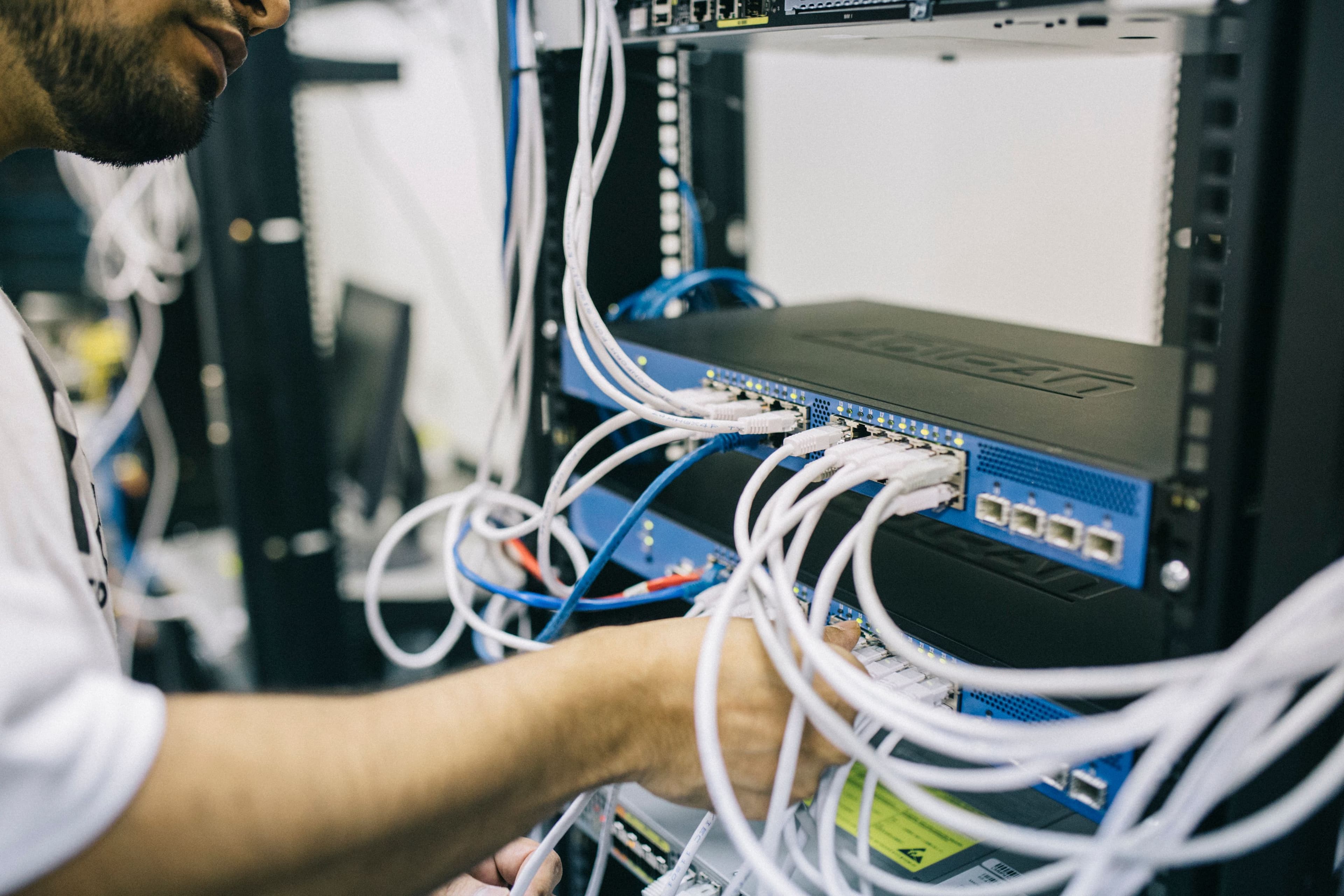 A technician in a white T-shirt dressing white Ethernet cables into a blue network switch mounted in a black rack, green link lights on the ports. — Photo: Field Engineer, "Electronics engineer fixing cables on server", via Pexels, Pexels License (https://www.pexels.com/license/)