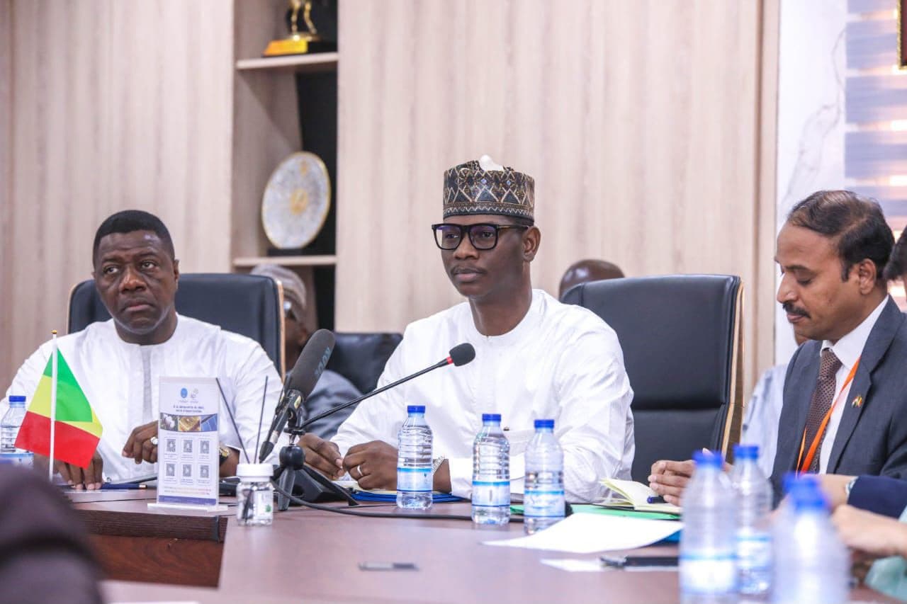 Meeting around a table fitted with microphones: at the centre a man in a white boubou and embroidered hat, to his right a man in a white boubou behind a small table flag, to his left a man in a suit taking notes, water bottles on the table.